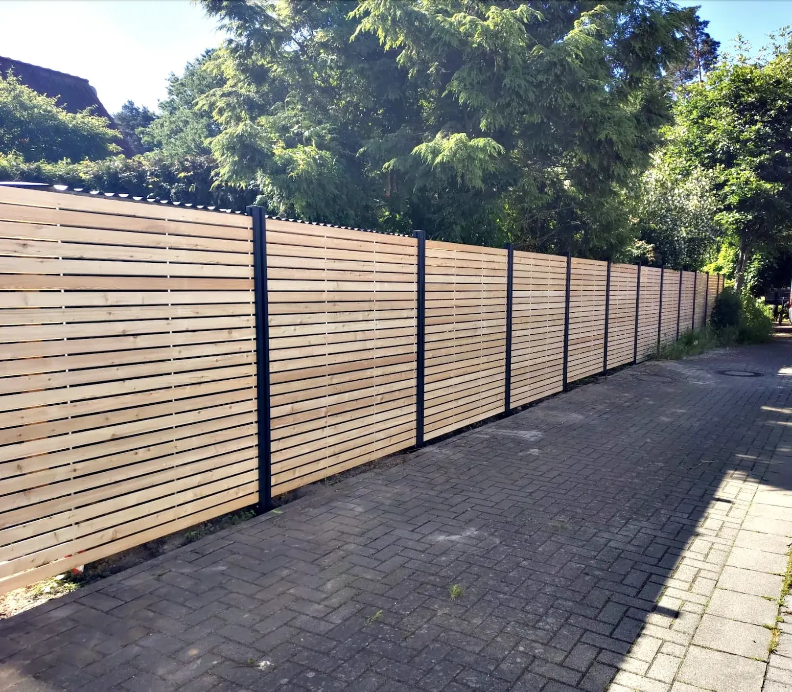 Langer Rhombuszaun mit dunklen Pfosten an einer gepflasterten Zufahrt